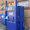 Outside, against a brick wall stands the BP18 bale press. Its door and panels are closed and locked. A blue wheelie bin can be seen behind to the left.