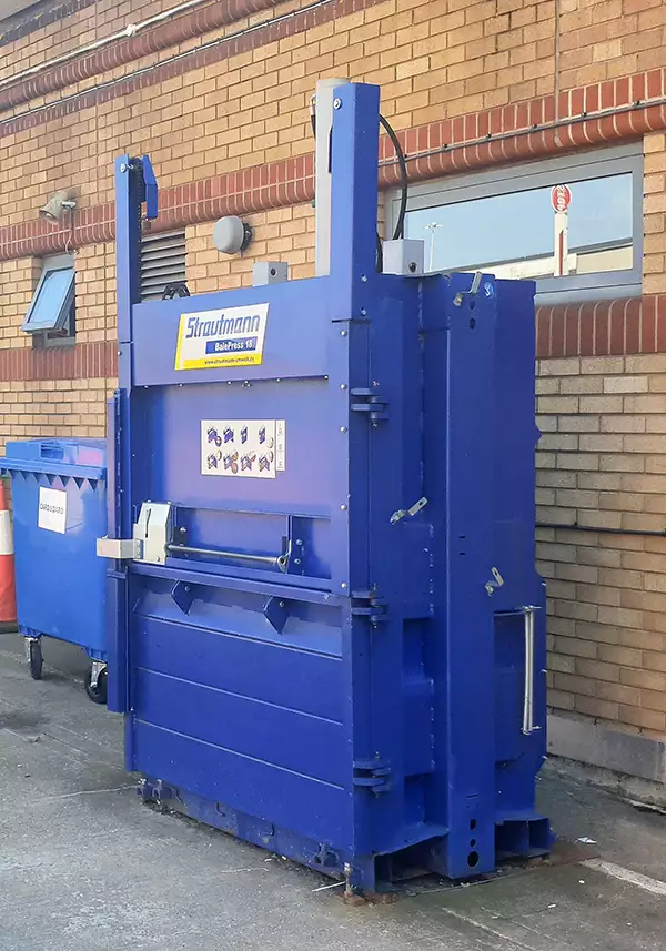 Outside, against a brick wall stands the BP18 bale press. Its door and panels are closed and locked. A blue wheelie bin can be seen behind to the left.