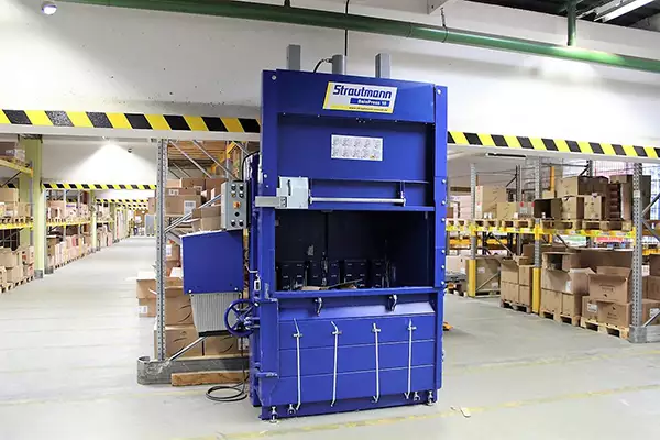 The blue BP18 baler stands in a warehouse with its front compartment open. A small amount of material can be seen within the baler. In the background, cardboard boxes are stacked on shelves.