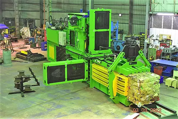 A green and yellow CB-L Channel Baler stands in the middle of a large warehouse. A compacted bale of carboard can be seen breaching the front exit of the baler.