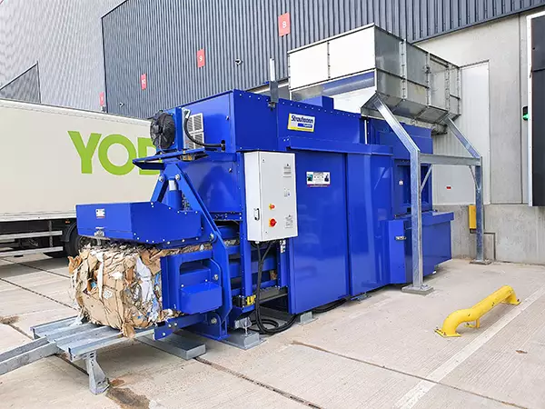 Outside a warehouse, the GigaBaler stands with a large bale of compacted cardboard protruding from its front opening. A large metal structure stands astride the baler at the back for entry of cardboard.
