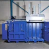 Front view of the MK1100 Twin baler in blue with the right-side door ajar. Nothing is inside the machine. On top to the right sits the large grey device with three protruding rods.