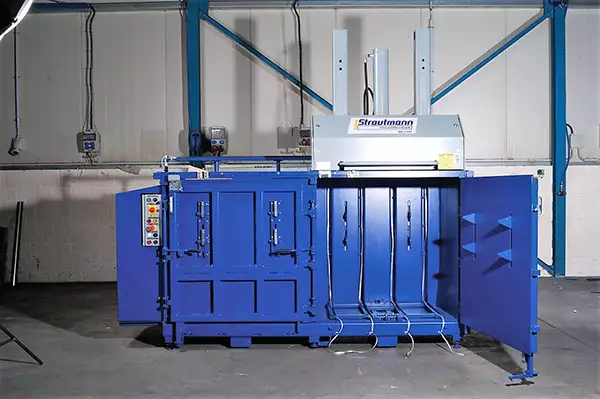 Front view of the MK1100 Twin baler in blue with the right-side door ajar. Nothing is inside the machine. On top to the right sits the large grey device with three protruding rods.