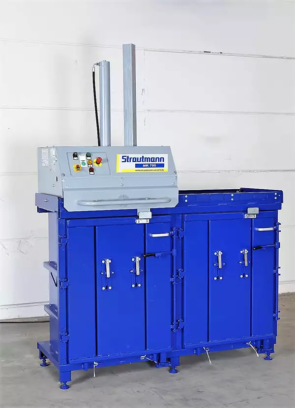 Angled front view of the MK700 Twin baler which stands against a white wall. Atop the baler to the left is a grey device adorned with coloured buttons.