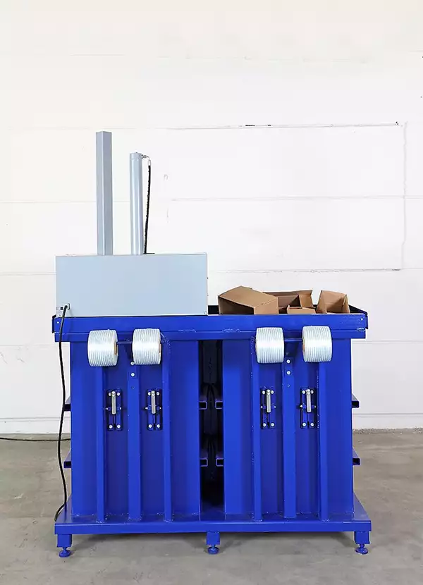 The back of the blue MK700 shows four rolls of white tape or cord, two on either side of the twin machine. Atop the machine to the left, the back of a grey device can be seen with cardboard protruding from inside at the right.