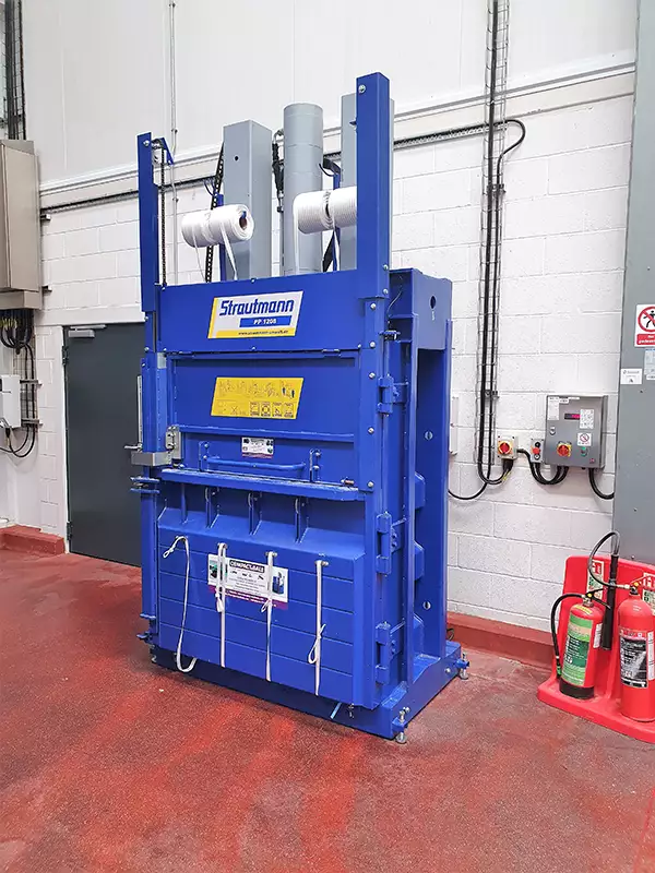 The PP1208 baler stands against a warehouse wall on a red floor, contrasting the blue of the machine.