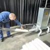 A man stands bent over as he pulls compacted Styrofoam tubes from the Styropactor Nano. The Styropactor Nano has four curved legs and a sticker of two eyes on the front of its metal cuboid shell.