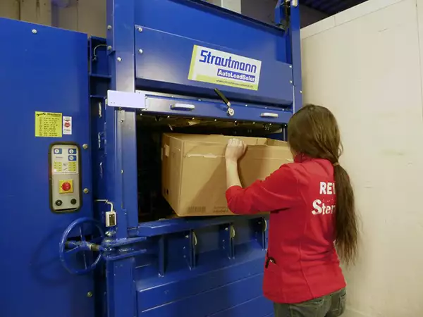 A woman faces away while loading a cardboard box into the blue Auto Load Baler. She is wearing a red jumper with lettering obscured by her ponytail on the back.