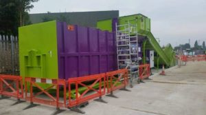 A large green and purple food compactor stands outside with scaffolding and safety fences around it.